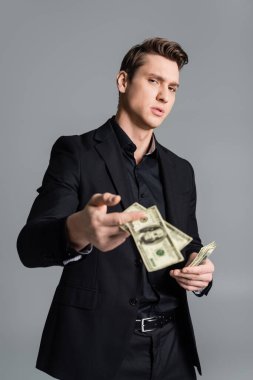 confident man in formal wear showing blurred dollars at camera isolated on grey