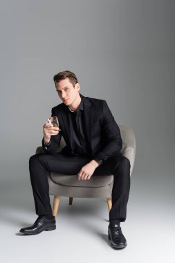 full length of stylish man with glass of whiskey sitting in armchair on grey background clipart