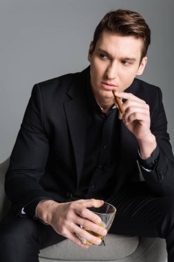 confident man in black holding cigar and glass of whiskey while looking away isolated on grey clipart