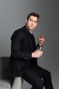serious man in black blazer looking away while sitting with whiskey and cigar isolated on grey