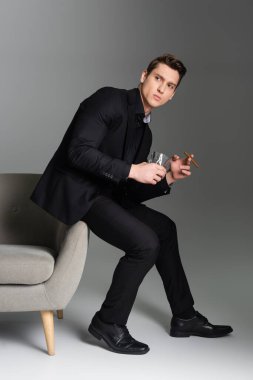 Viski ve puro içen bir adam koltukta oturuyor ve gri arka plana bakıyor.