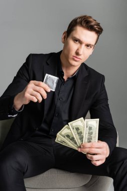 confident man holding condom and dollars while looking at camera isolated on grey clipart