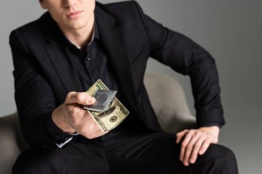 cropped view of blurred man holding condom and dollar banknote isolated on grey clipart