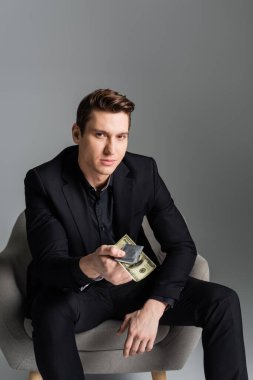 man in black suit holding condom and money while looking at camera isolated on grey