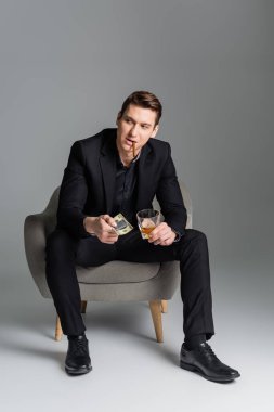Gri arka planda otururken elinde dolar ve prezervatif tutan puro ve viskiyle tam boy bir adam.