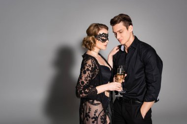 woman in lace mask and tunic clinking champagne glasses with man on grey background