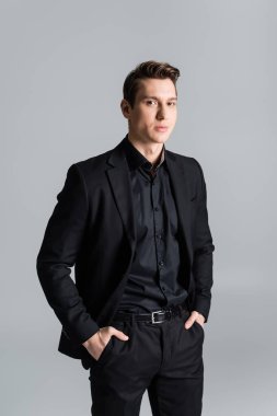 confident man in black suit standing with hands in pockets and looking at camera isolated on grey