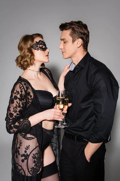 side view of man in black shirt and woman in lace mask clinking champagne glasses on grey background
