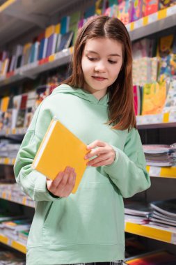 Çocuk kırtasiyedeki bulanık rafın yanındaki yeni deftere bakıyor.