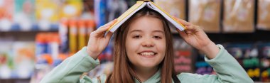 cheerful girl covering head with copybook and looking at camera in blurred stationery shop, banner