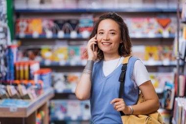 cheerful student with backpack talking on smartphone in blurred stationery shop
