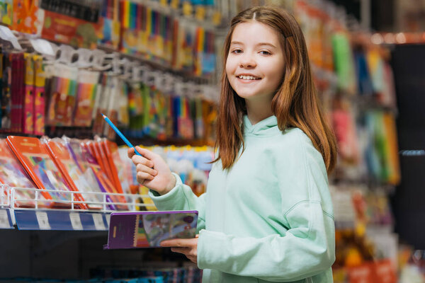 joyful girl with pencil set smiling at camera in stationery shop