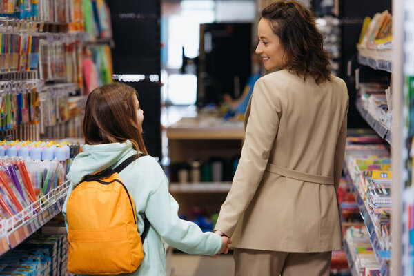 back view of smiling woman and girl with backpack holding hands in stationery store