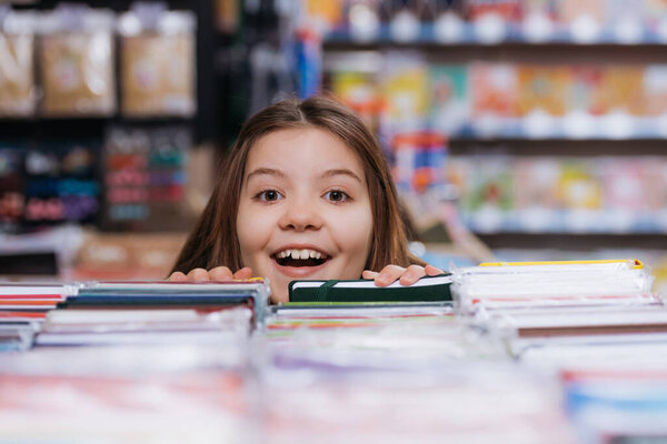excited child looking at camera near blurred copybooks in stationery store