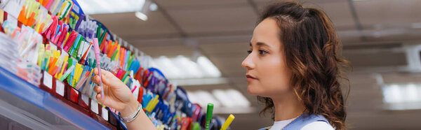 young brunette woman choosing pen on rack in stationery store, banner
