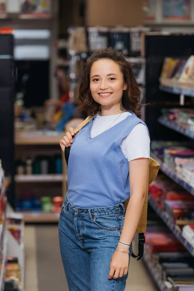 cheerful student with backpack looking at camera in stationery store