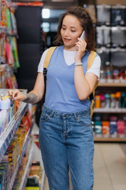 tattooed woman talking on mobile phone near rack in stationery shop