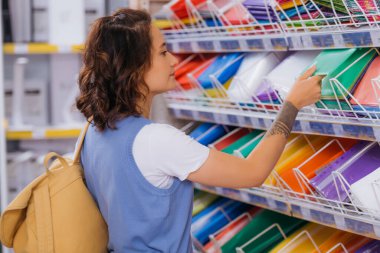 brunette woman with backpack choosing plastic files in stationery store