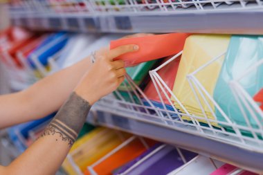partial view of tattooed woman taking plastic folder from rack in stationery shop clipart