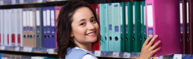 happy woman taking folder from rack in stationery store and looking at camera, banner