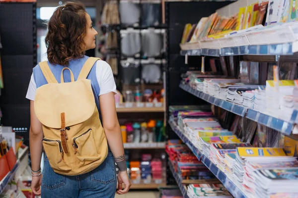 back view of woman with backpack looking at rack with copybooks in stationery store