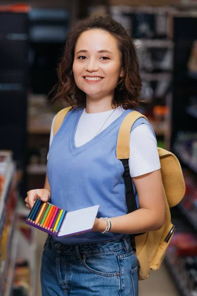 joyful woman with set of color pencils looking at camera in stationery shop