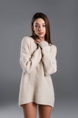 pretty woman in soft sweater touching neck and looking at camera on grey background