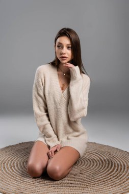 pretty woman in sweater sitting on rug with naked legs and looking away on grey background clipart