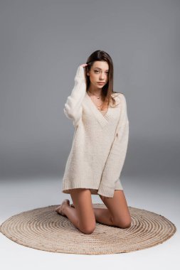 young woman in sweater and with bare legs kneeling on carpet on grey background