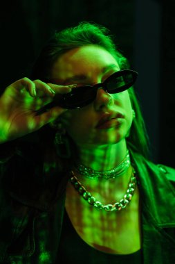 portrait of trendy woman in silver accessories adjusting sunglasses in green neon light isolated on black