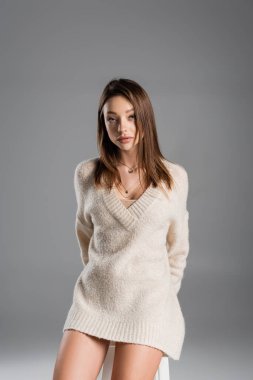 young woman in cozy sweater standing with hands behind back and looking at camera on grey background