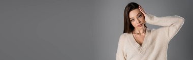 brunette woman in cozy sweater touching head and looking at camera isolated on grey, banner