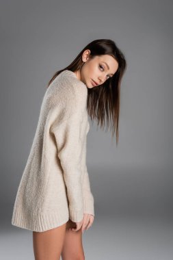 brunette woman in long and soft sweater looking at camera on grey background clipart