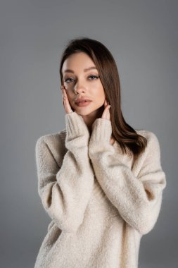pretty woman in cozy sweater touching face while looking at camera isolated on grey