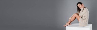 brunette woman with naked legs sitting on white cube and looking at camera isolated on grey, banner