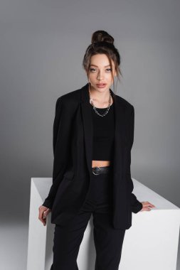 brunette woman in black suit and silver necklace looking at camera near white cube on grey background