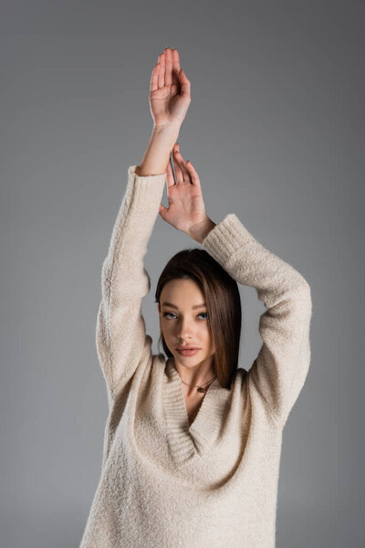 sensual woman in warm sweater standing with raised hands and looking at camera isolated on grey