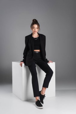 full length of woman in black sneakers and suit sitting on white cube on grey background