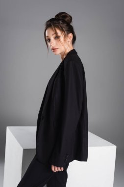 young and stylish woman with makeup standing in black blazer near white cube isolated on grey