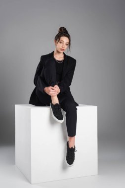 full length of woman in black suit and sneakers sitting on white cube on grey background