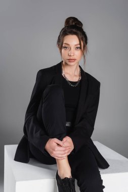 charming woman in black jacket sitting on white cube and looking at camera isolated on grey