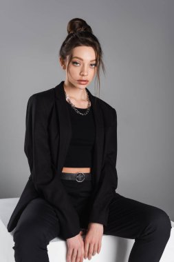 pretty woman with makeup sitting in black clothes on white cube isolated on grey