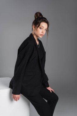 young and trendy woman in black blazer looking at camera near white cube isolated on grey
