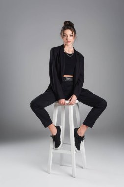full length of brunette woman in black clothing sitting on white stool on grey background