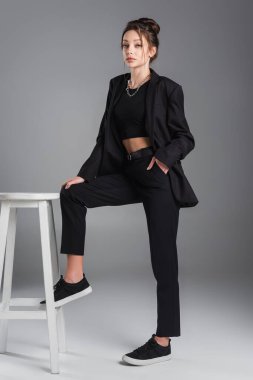 young brunette woman posing with hand in pocket of black trousers near white stool on grey background