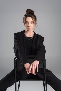 brunette woman in black clothes sitting on chair and looking at camera isolated on grey