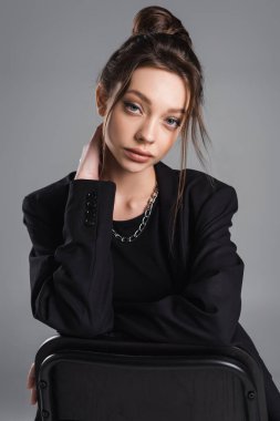 charming woman in black blazer touching neck while looking at camera isolated on grey