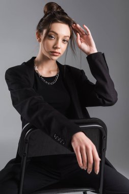 young woman in black jacket and metal necklace fixing hair while sitting isolated on grey