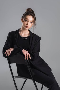 sensual woman in black suit sitting with crossed arms on chair isolated on grey