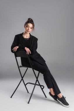 full length of woman in black stylish suit and sneakers posing on stool on grey background